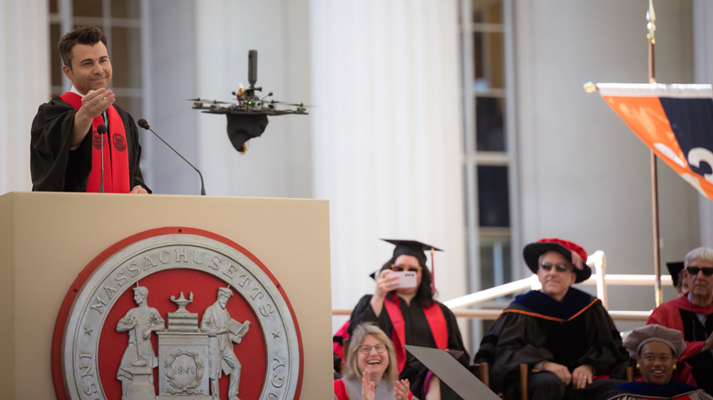 Mark Rober gestures to a drone, which is flying up with his graduation cap